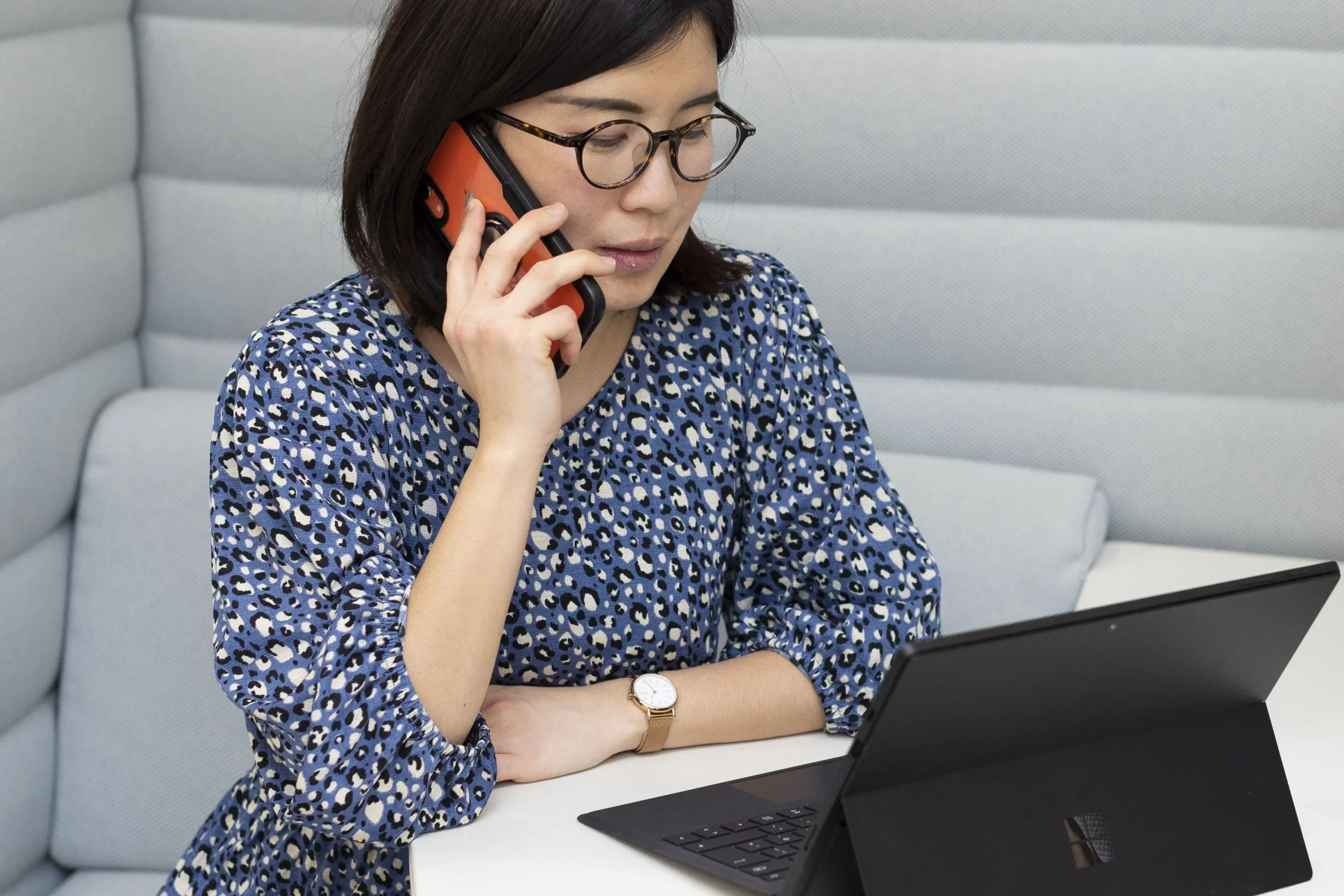 A woman in a blue printed dress talks on her phone while looking at a laptop in a cozy booth. She appears focused, creating a professional tone.