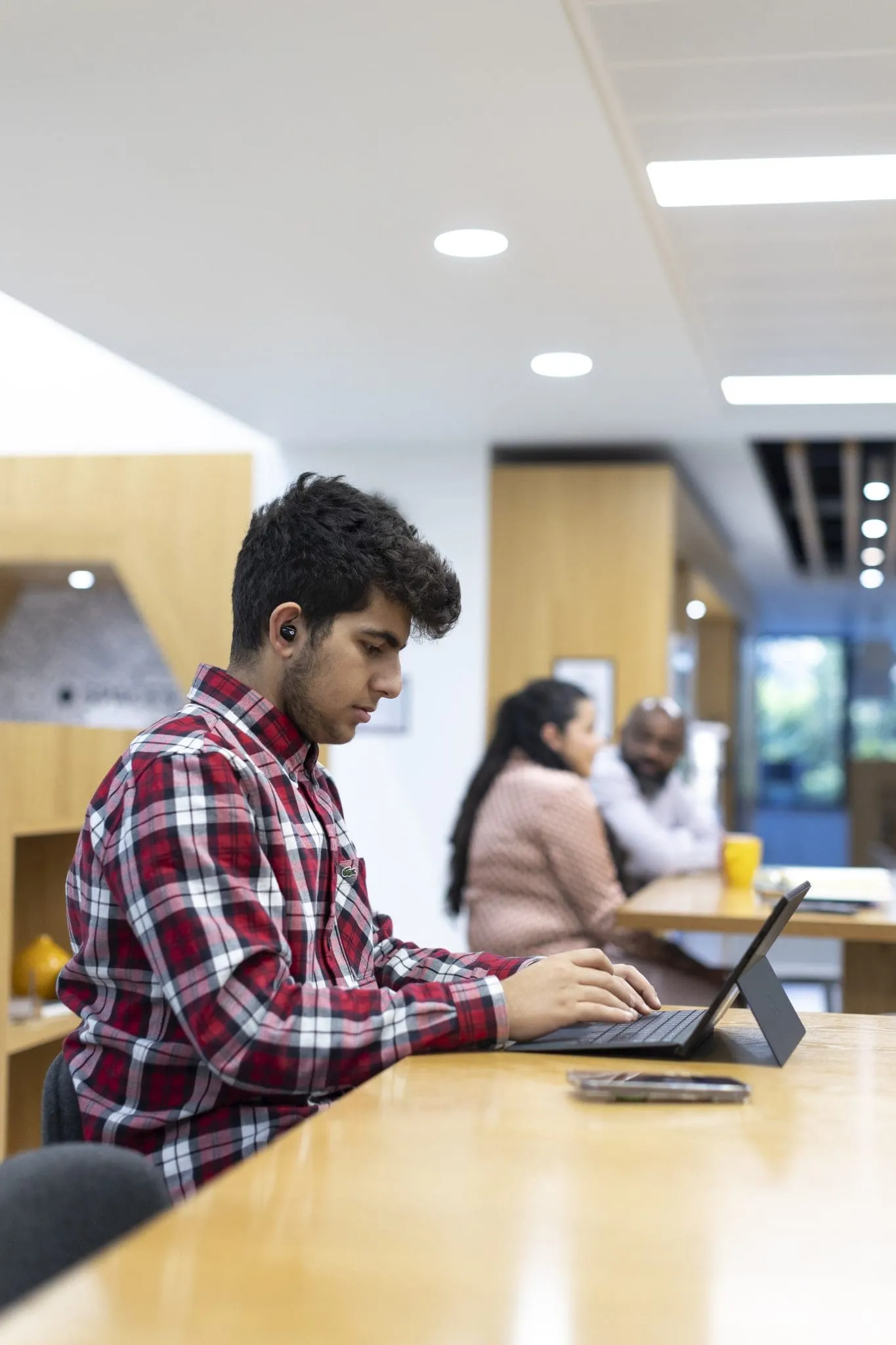 A young man in a red plaid shirt works on a laptop at a wooden table in a modern office, wearing earbuds. Two colleagues chat in the blurred background.