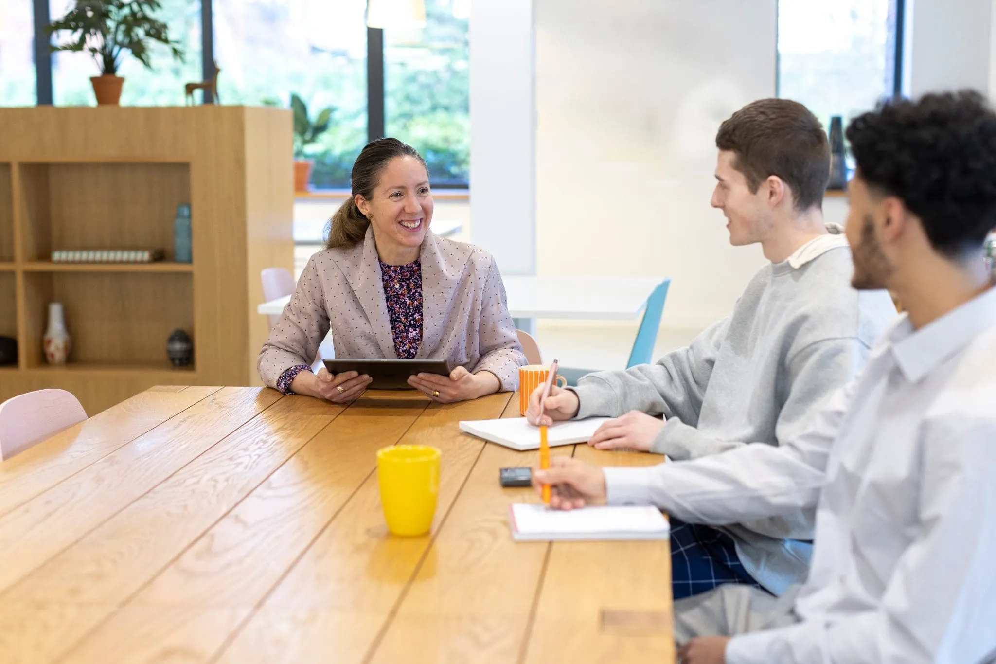 A woman with a tablet smiles while speaking to two men seated at a table in a bright office. They have notebooks and mugs, creating a collaborative mood.