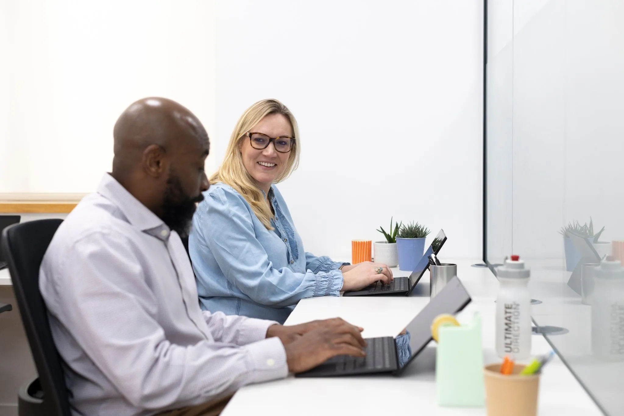 Two colleagues sit side-by-side at a desk with laptops. The woman smiles at the man. Desk items include colorful mugs and potted plants, conveying a friendly office vibe.