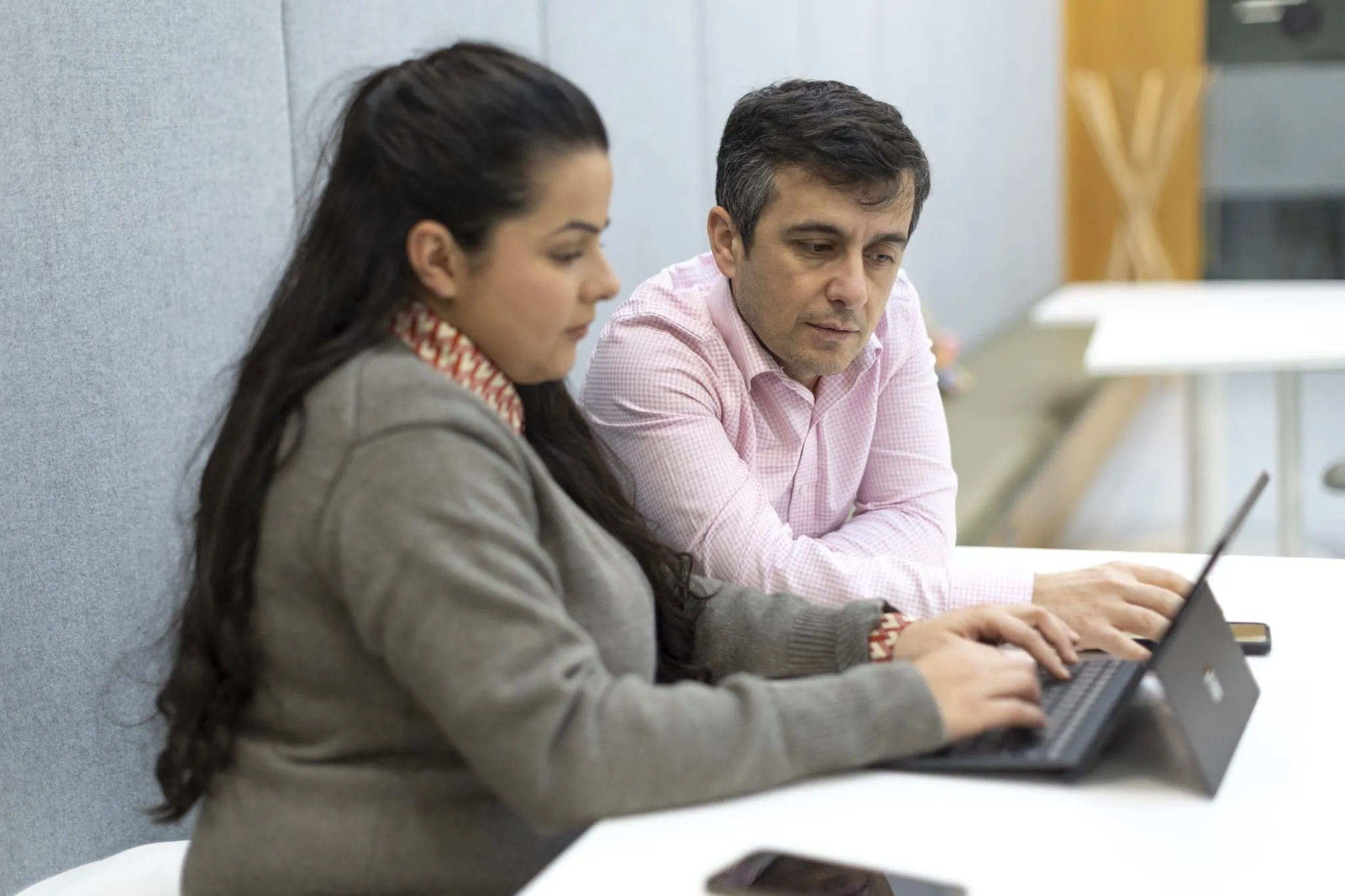 Two people are seated at a desk in a modern office, focused on a laptop. The scene conveys a collaborative and professional atmosphere.